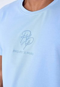 Camiseta de algodón azul claro con un logo bordado y el texto "PROJECT X PARIS" en un azul más oscuro. Vista cercana de la textura de la tela.