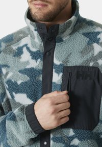 Sudadera de forro polar en patrón de camuflaje azul y blanco. Presenta un bolsillo con cremallera negra y cierre de botón en el cuello. Textura suave.