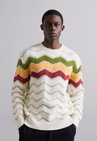 Balts adīts džemperis ar zigzaga rakstu zaļā, oranžā, sarkanā un bēšā krāsā. Tam ir ribota apkakle un sānu daļas.