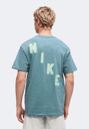 Personne blonde portant un T-shirt bleu sarcelle à manches courtes avec de grandes lettres vert clair formant "NIKE" en diagonale dans le dos.