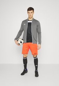 Veste de sport zippée grise, t-shirt noir, shorts orange vif avec accents noirs, chaussettes noires et chaussures de football noires. Tenant un ballon de football.