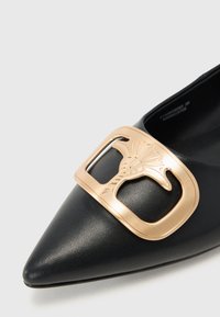 Chaussure noire en cuir à bout pointu, dotée d'une boucle dorée proéminente avec un design décoratif. Texture lisse et silhouette élégante.