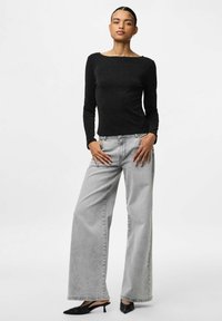 Schwarzes langärmeliges Oberteil mit einem weiten Ausschnitt, kombiniert mit hellgrauen weit geschnittenen Jeans. Das Model trägt schwarze spitz zulaufende Pumps. Minimalistisches Design.