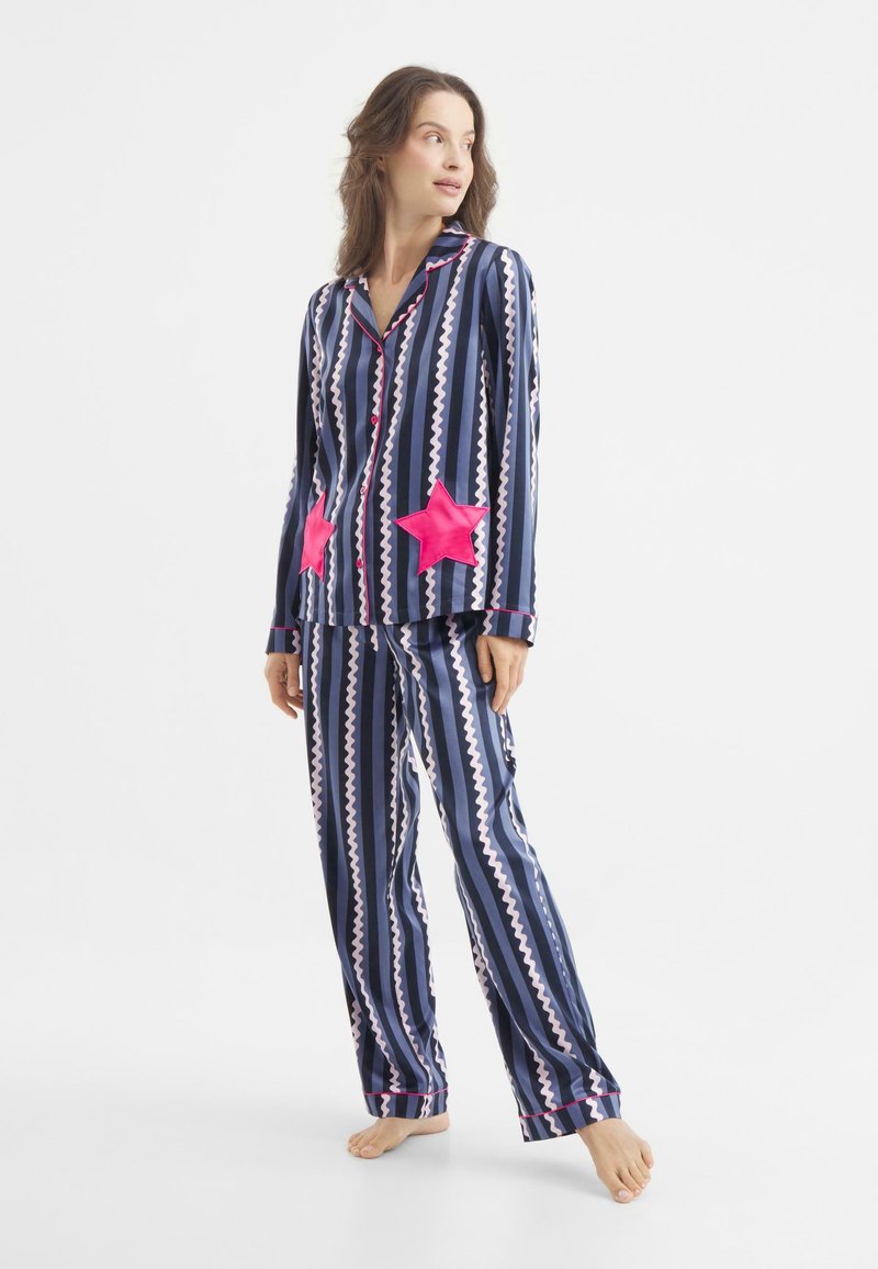 Navyblauw gestreepte pyjama set met roze zigzagpatronen en felroze sterapplicaties op de voorzakken. Zachte stof, lange mouwen, losse pasvorm.