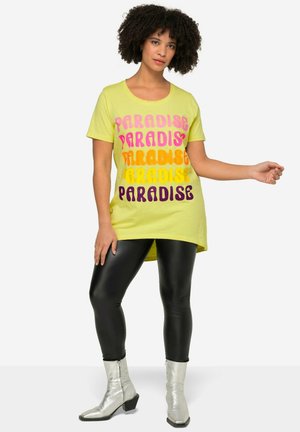 Gul t-shirt med flerfarvet "PARADISE" tekst i pink, orange og lilla; sat sammen med sorte, skinnende leggings og sølvfarvede ankelstøvler.