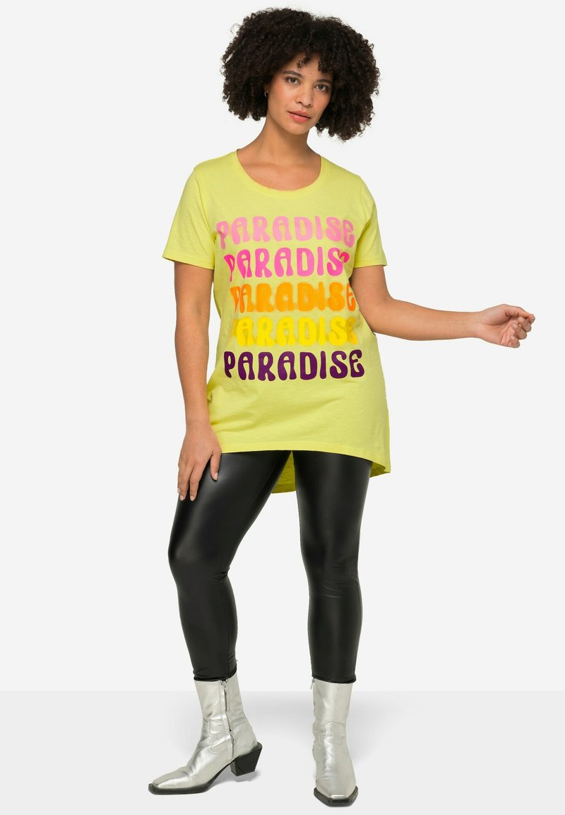 T-shirt gialla con testo "PARADISE" multicolore in rosa, arancio e viola; abbinata a leggings neri lucidi e stivaletti argentati.