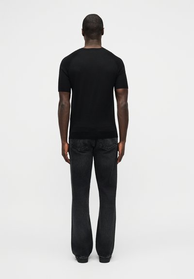 Άνδρας που στέκεται με την πλάτη, φοράει εφαρμοστή μαύρη κοντομάνικη μπλούζα και χαλαρά μαύρα τζιν, μπροστά σε απλό λευκό φόντο.