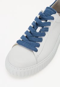 Bílá kožená teniska s kulatou špičkou, s modrými denimovými tkaničkami a texturovanou gumovou podrážkou. Minimalistický design s čistými liniemi.