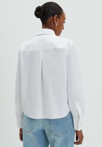 White shirt made of smooth fabric, featuring a back pleat, long sleeves, and buttoned cuffs. Paired with light blue denim jeans.