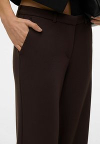 Bruna skräddarsydda byxor med en slät textur, sidofickor och en figursydd midjeresår, visas med en hand placerad i en ficka.