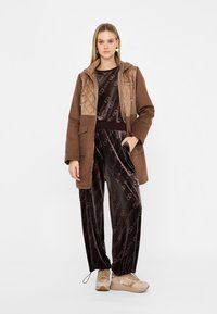 Giacca marrone trapuntata con cappuccio, indossata sopra un completo di felpa nera in velluto con un motivo a logo. Sneakers beige completano l'outfit.