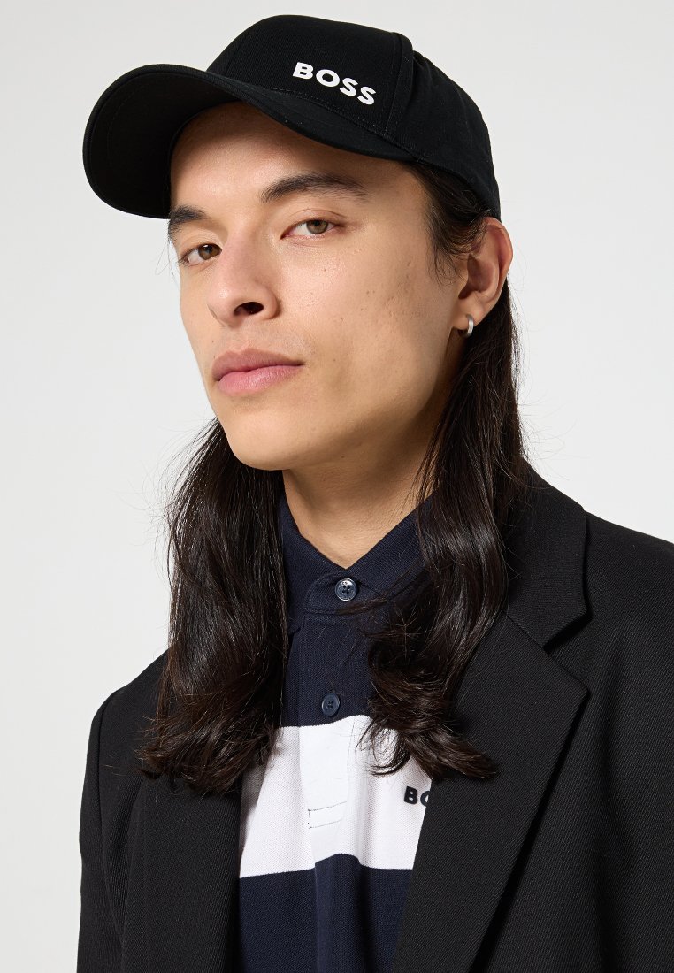 Junge Person mit langen dunklen Haaren, die eine schwarze "BOSS"-Kappe, ein schwarzes Blazer, ein gestreiftes Poloshirt und einen kleinen Creolenohrring trägt, vor einem schlichten Hintergrund.