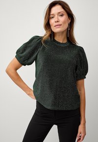 Groen sprankelend topje met korte puffmouwen, een hoge halslijn en een ontspannen pasvorm. G gedragen met zwarte broeken, met een gladde textuur.