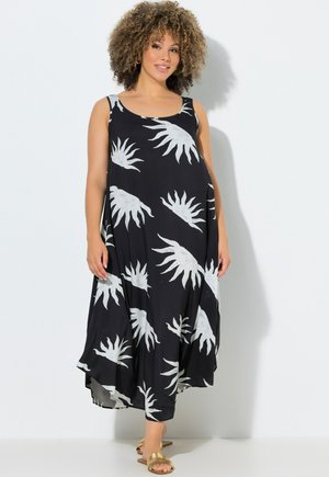 Musta hihaton mekko, jossa valkoiset lehtikuvioinnit, virtaava malli, kevyt kangas ja epäsymmetrinen helma. Malli käyttää kultaisia sandaaleja.