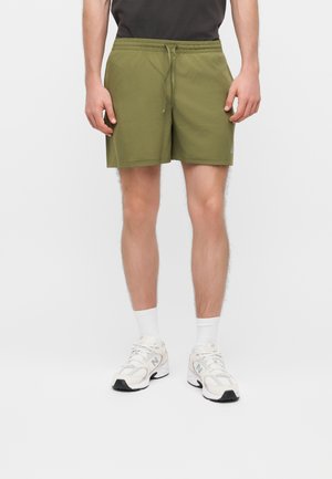 Mann trägt olivgrüne Shorts mit Kordelzug, weiße Socken und weiße Sportschuhe, steht vor einem einfarbigen hellen Hintergrund.