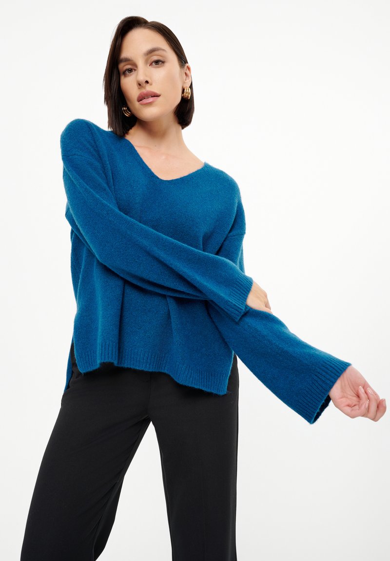 Turkusowy, oversize sweter z dekoltem w kształcie litery V, długimi rękawami i bocznymi rozcięciami. Wykonany z miękkiego, strukturalnego materiału, w połączeniu z czarnymi spodniami.