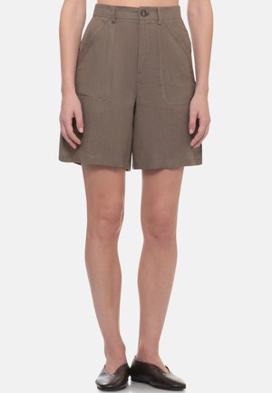 Donna che indossa pantaloncini marroni a vita alta fino al ginocchio con tasche frontali e chiusura con bottoni, abbinati a scarpe basse nere.