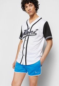 Hvit baseball-lignende trøye med svarte ermer, med en stor svart skriptlogo og knapplukking. Kombinert med blå shorts med hvit kant.