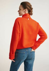 Jasnopomarańczowy sweter z dzianiny z wysokim kołnierzem i ściągaczami, połączony z niebieskimi dżinsami, z detalem guzików z tyłu.