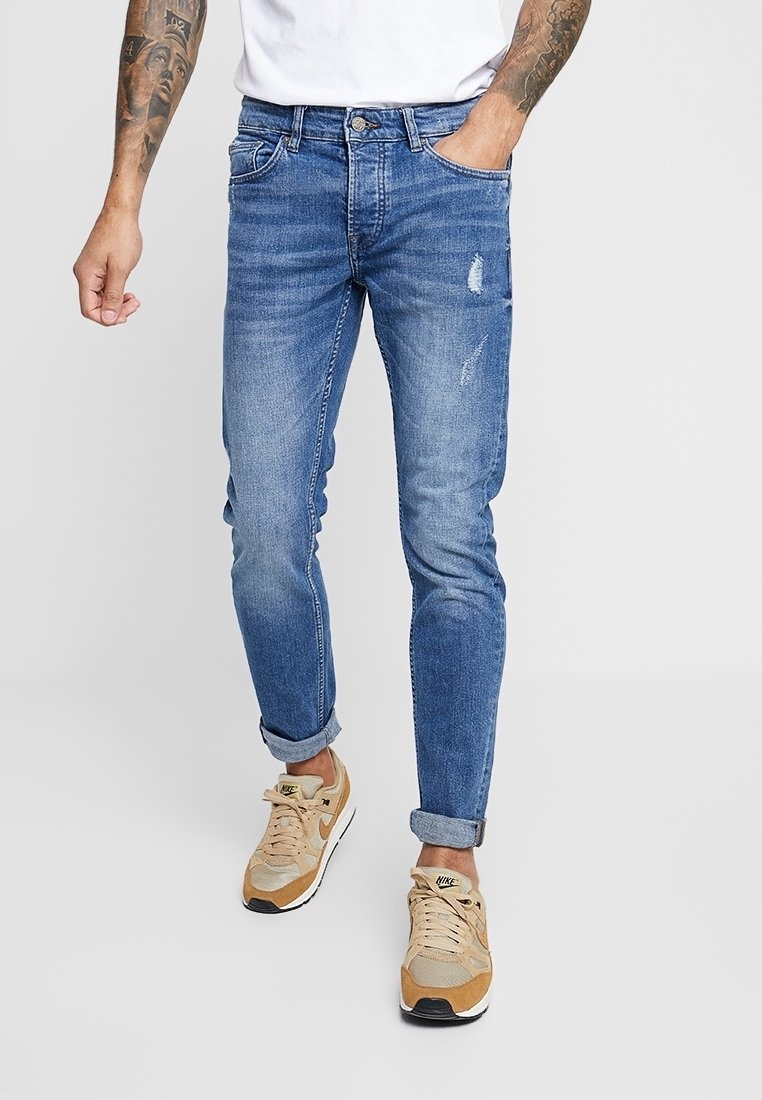 Ljusblå denimjeans med smal passform, med subtila slitningar och uppvikta benslut, kombinerade med beigea sneakers.