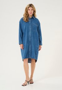 Denim-paita-mekko keskisinisessä värissä, jossa on pitkät hihat, nappikiinnitys, rintatasku ja korkea-matala helma. Tyylitettu mustilla remmikengillä.