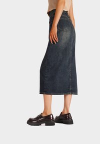 Gonna midi in denim blu scuro con taglio dritto, caratterizzata da tasche posteriori e cuciture a contrasto. Indossata con mocassini scuri.