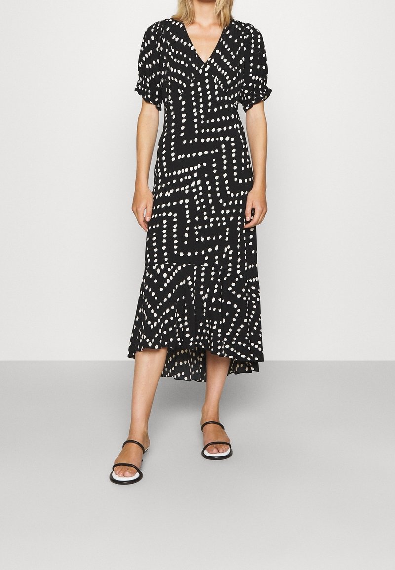 Femme portant une robe midi noire avec un motif en zigzag de points blancs irréguliers, des manches bouffantes et des sandales noires à bouts ouverts avec des fines lanières.