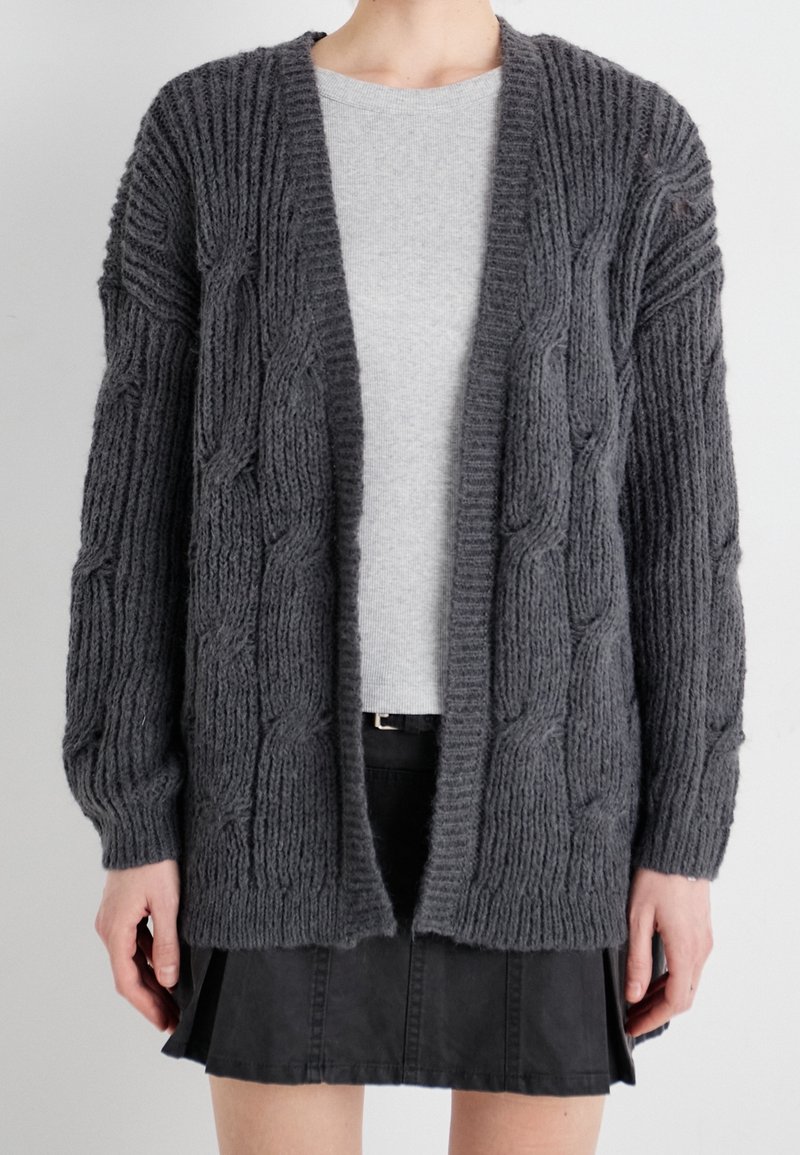 Personne portant un cardigan en maille torsadée épaisse gris foncé sur un haut gris clair et une jupe plissée noire, sur un fond uni.