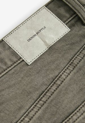 Grau denim Stoff mit vertikalen Nähten und einem rechteckigen Lederetikett mit der Aufschrift "DENIM SUPPLY", das mit dunklem Faden befestigt ist.