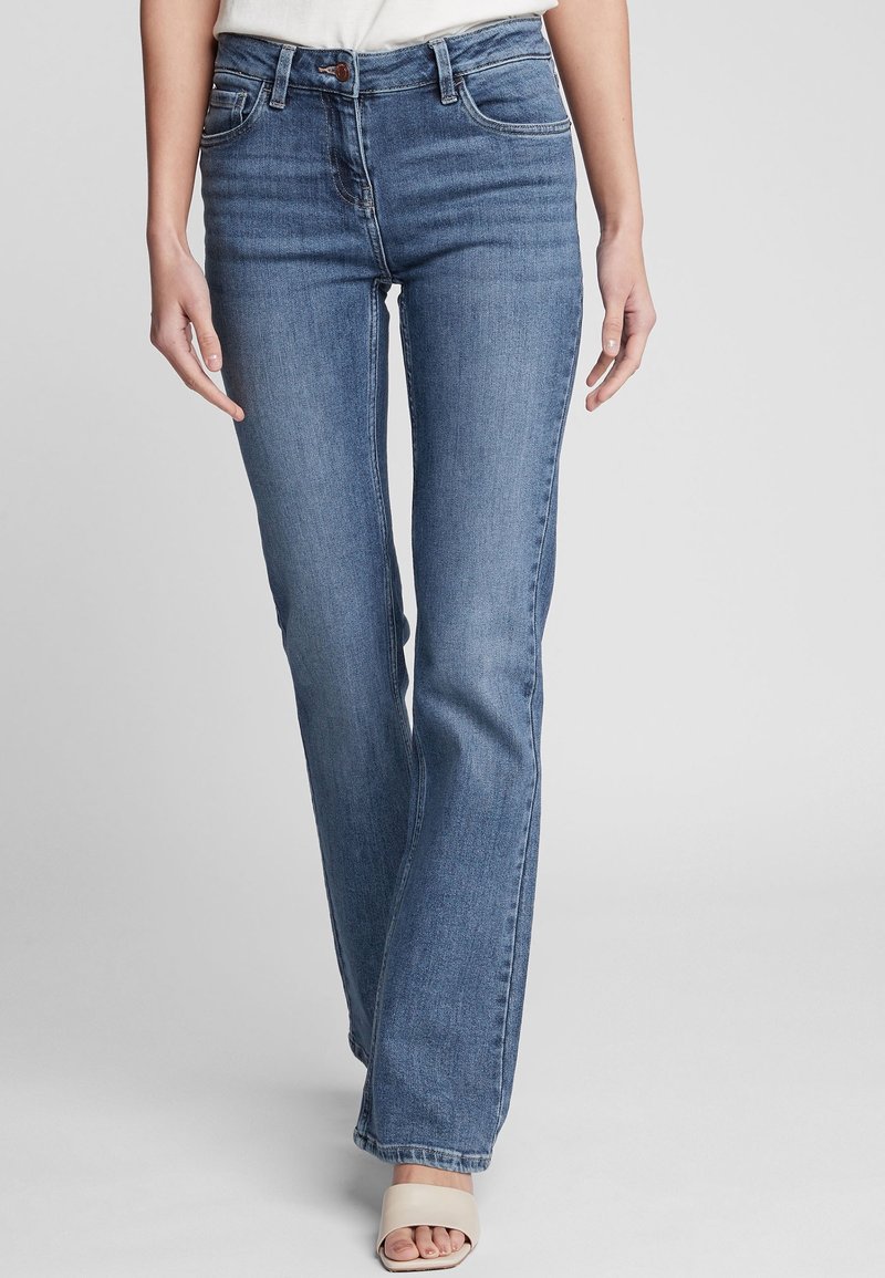 Femme portant un jean bootcut en denim bleu taille mi-haute et des sandales blanches à talons ouverts, debout devant un fond uni gris clair.