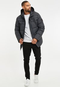 Grå puffjacka med huva, dragkedjefickor och en logopatch, ihop med svarta byxor och vita sneakers. Slät textur, isolerad design.