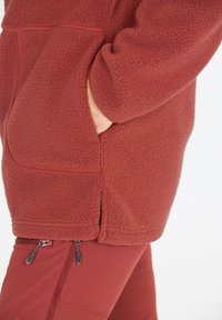 Rote Fleecejacke mit strukturiertem Oberflächenmaterial, Seitentaschen und lockerer Passform, kombiniert mit passenden roten Hosen mit Reißverschlussakzenten.