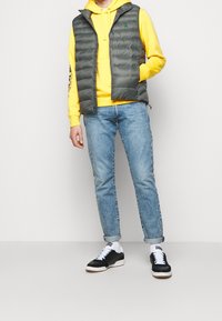 Herrarnas gråa pufferväst över en gul hoodie, ihop med ljusblå jeans och svarta sneakers. Västen har en dragkedja och en logotyp.