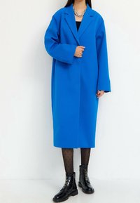 Abrigo extragrande de mezcla de lana azul con solapa, botonadura frontal y mangas largas, combinado con medias negras con lunares y botas de tobillo brillantes.