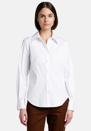 Camisa blanca de botonadura hecha de algodón, con cuello tipo pico, mangas largas y dobladillo curvado. Combinada con pantalones de sastre marrones.