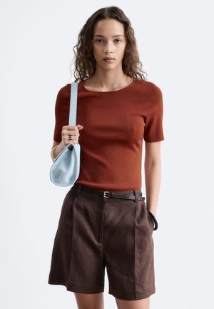 Femme aux cheveux bouclés portant un top manches courtes couleur rouille et un short marron taille haute, tenant un sac à main bleu clair sur son épaule.
