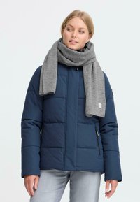 Kvinde iført en navy kviltet jakke, lysegrå jeans og et tykt gråt tørklæde, der ser til siden mod en ensfarvet lys baggrund.