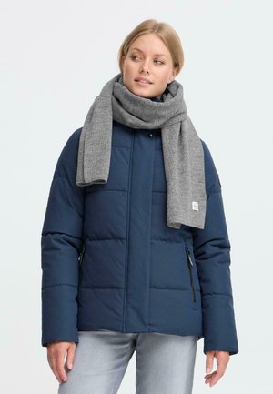 Kvinne iført en marineblå quiltet jakke, lysegrå jeans og et tykt grått skjerf, som ser mot siden mot en enkel lys bakgrunn.