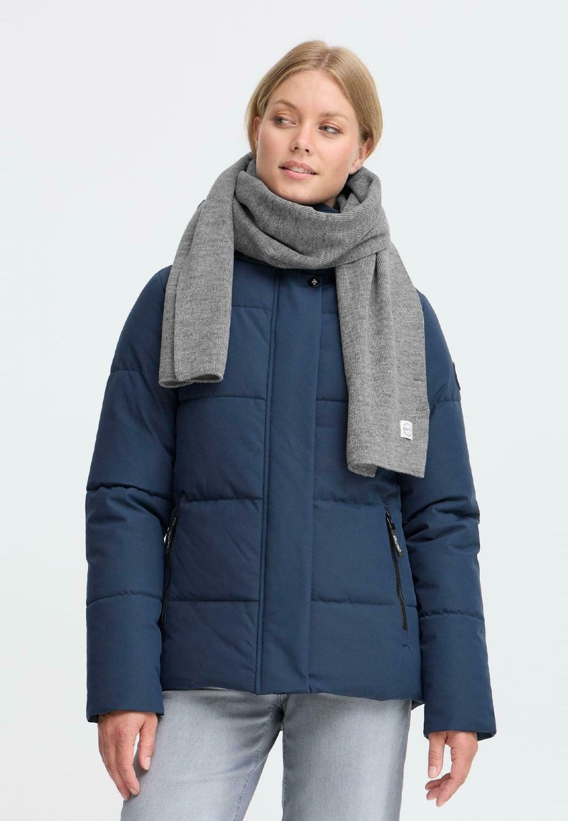 Kvinde iført en navy kviltet jakke, lysegrå jeans og et tykt gråt tørklæde, der ser til siden mod en ensfarvet lys baggrund.