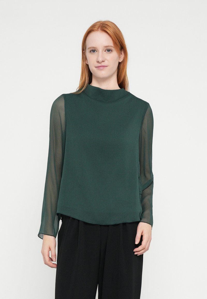 Tmavozelená blúzka s vysokým, zaobleným výstrihom. Hlavná látka je jednoliata, zatiaľ čo dlhé rukávy sú priehľadné. Hladká textúra, pohodlný strih.