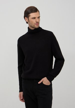 Maglione a collo alto di colore nero realizzato in un morbido tessuto a maglia, con polsini e orlo a costine, abbinato a pantaloni neri, in piedi di fronte a uno sfondo semplice.