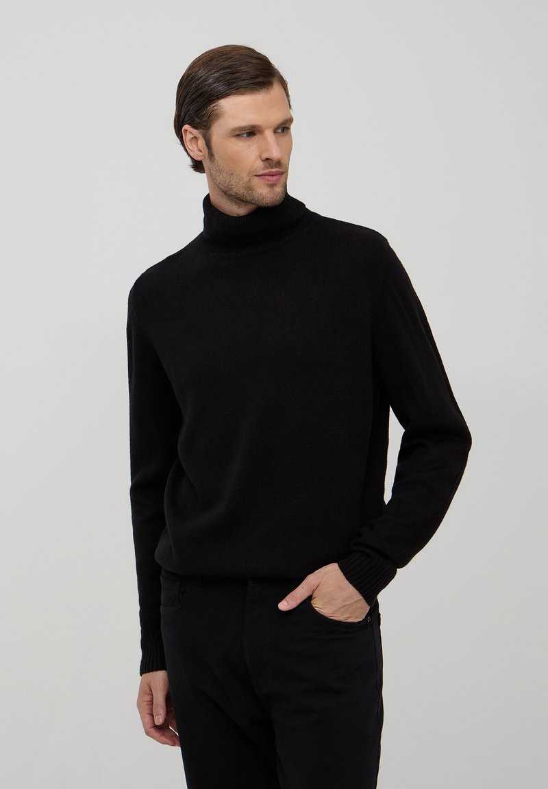 Maglione a collo alto di colore nero realizzato in un morbido tessuto a maglia, con polsini e orlo a costine, abbinato a pantaloni neri, in piedi di fronte a uno sfondo semplice.