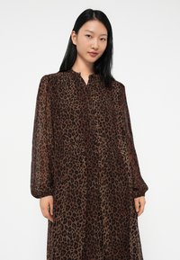 Jauna sieviete ar gariem melniem matiem, ģērbusies brūnas un melnas leoparda raksta kleitā ar garām piedurknēm, uz viendabīga baltā fona.