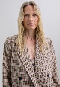 Checkered blazer in soft fabric, featuring a double-breasted design, muted pink and brown tones, with a V-neck underneath and prominent collar.