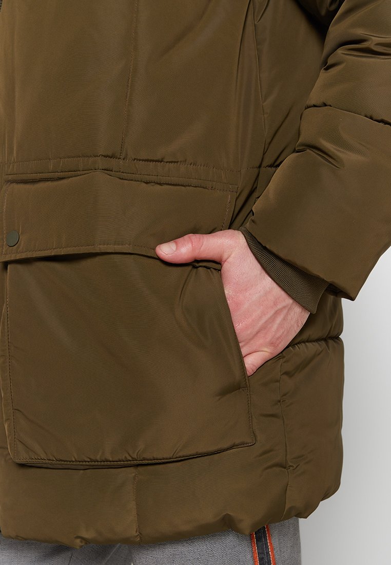 Olivgrüne Steppjacke aus strukturierter Stoff, mit großer Fronttasche und einer Nahaufnahme einer Hand, die in der Tasche ruht.