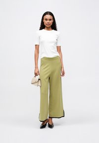 Biały top z krótkim rękawem w połączeniu z szerokimi spodniami w kolorze szałwii z czarnym wykończeniem, trzymająca małą beżową torebkę o strukturalnym wzorze.