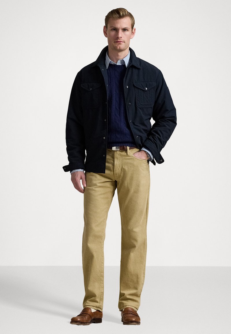 Marinejacke mit zwei Vorderaschen über einem dunkelblauen Strukturstrickpullover, kombiniert mit khakifarbenen Hosen und braunen Slippern.