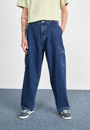 Personne portant un pantalon cargo en denim bleu foncé ample et des baskets grises, debout sur un tapis bleu avec une main dans la poche.