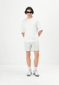 Camiseta blanca de algodón y pantalones cortos de cuadros en azul claro. El modelo lleva unas gafas de sol negras y zapatillas blancas con rayas negras.