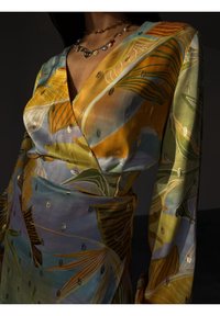 Femme portant une robe portefeuille en satin colorée avec des motifs de feuilles jaunes, vertes et oranges et un collier pendentif en or sur un fond sombre.